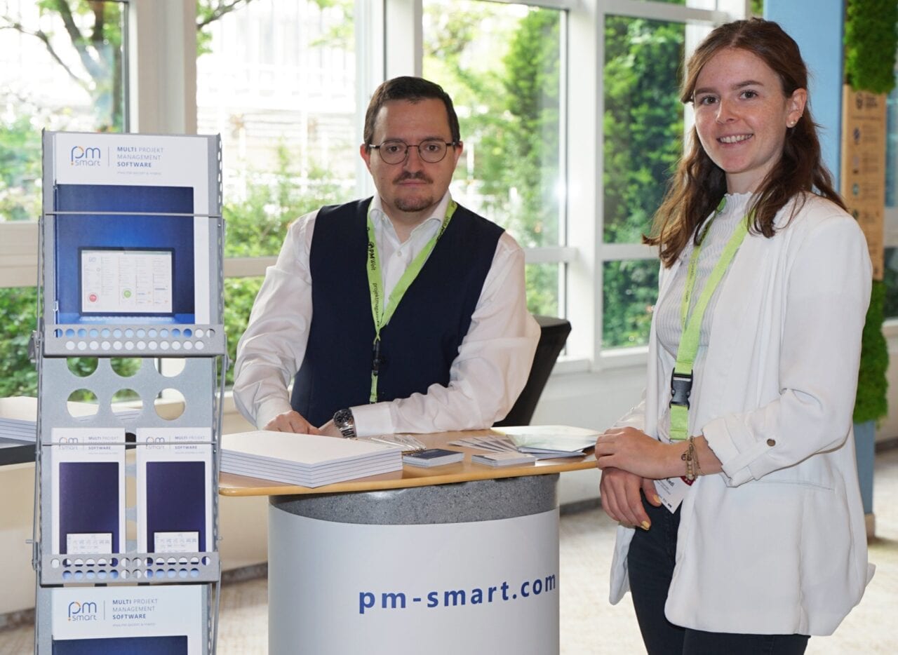 Auf dem Bild sind zwei Kolleg:innen am Messestand auf der PMWelt in München zu sehen. Beide Lächeln in die Kamera und lehnen am Ausstellertisch, auf dem Broschüren und Flyer liegen.