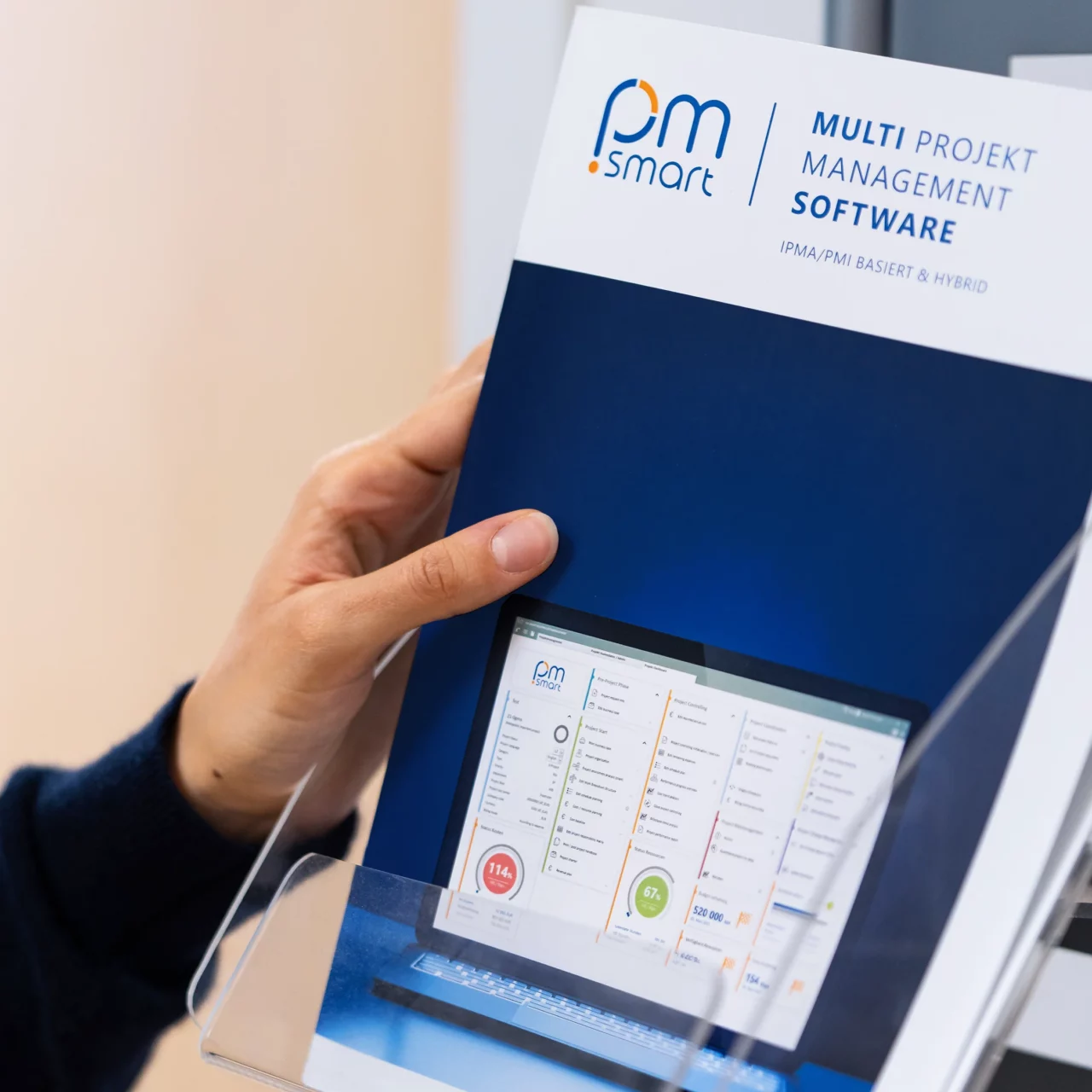 Foto von einer Hand, die von links eine Broschüre hält. Auf dem Cover ist das Logo der PM-Software pm-smart in Blau auf weißem Hintergrund abgebildet. Darunter befindet sich ein quadratisches, dunkelblau eingefärbtes Bild eines Laptops mit einer Projektmanagement-Dashboard-Ansicht der Software pm-smart, verschiedenen Widgets, einer Spiegelung auf dem Display sowie Monitorlichtreflexionen auf der Oberfläche, auf der der Laptop steht. Abgebildet in einem quadratischen Format.