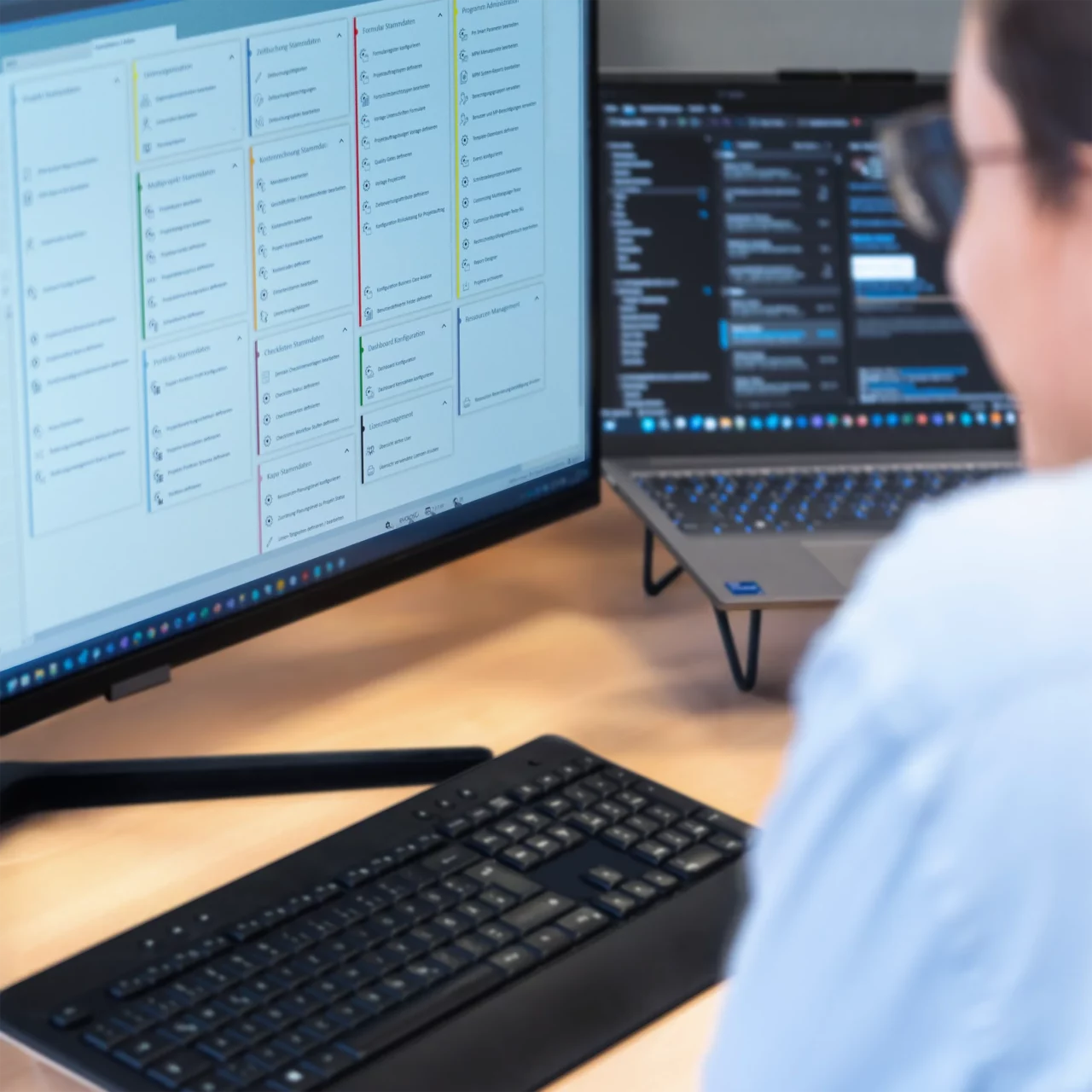 Dieses Foto zeigt einen Monitor mit einer Dashboard-Ansicht, der am linken Rand leicht angeschnitten ist und zahlreiche bunte Widgets enthält. Davor liegt eine schwarze Tastatur. Auf der rechten Seite des Bildes steht ein Laptop, der auf schwarzen Standfüßen ruht. Links im Vordergrund ist eine Person mit Brille und einem blauen Hemd zu sehen, deren Konturen jedoch verschwommen bleiben. Abgebildet in einem quadratischen Format.